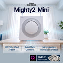 Coway Airmega Mighty2 Mini air purifier shown on a light wood table in a bright room, with large product text at the top and feature callouts along the bottom including IEST Certified HEPA, Quiet Mark Certified, and Microplastics Removal Certified.